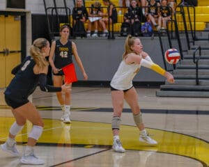 Volleyball player executes a forearm pass during a high school game, with teammates observing in the background.