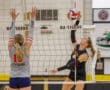 Volleyball player spikes ball over net against defender, intense game action.