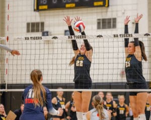Volleyball match action with NA Tigers players blocking at the net against Titans player preparing to hit the ball.