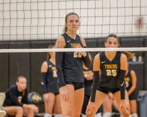 Volleyball players in action during a match, wearing black uniforms and focused behind the net.