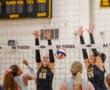 Volleyball match action: players attempt a block at the net while an opponent spikes the ball over the net.