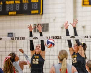 Volleyball match action: players attempt a block at the net while an opponent spikes the ball over the net.