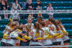 Volleyball team huddles on court, wearing white jerseys with yellow accents, against Pine-Richland Rams backdrop.