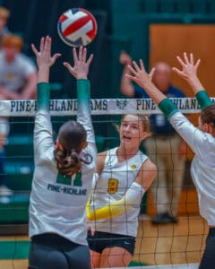 Female volleyball player spikes ball over net during intense indoor match.