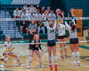 High school volleyball match with Pine-Richland players blocking a spike from the opposing team, crowd watching.