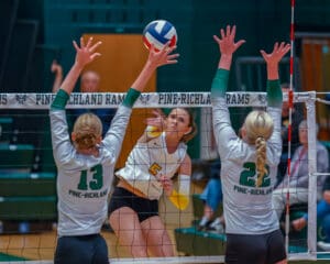 Volleyball player spikes ball past blockers during game. Pine-Richland Rams players attempt to defend.
