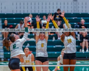 High school volleyball game action: player spikes ball as two opponents attempt block at Pine-Richland court.