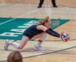 Volleyball player dives to save a ball during an indoor match on a wooden court.