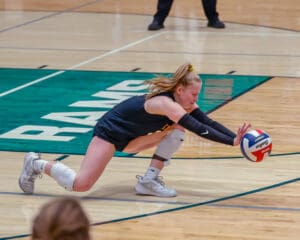 Volleyball player dives to save a ball during an indoor match on a wooden court.