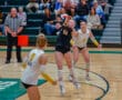 Women's volleyball game action, player in black jersey hitting the ball, opposing team players ready on the court.