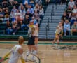 Women's volleyball match, player sets ball while teammates and crowd watch intensely in gymnasium.