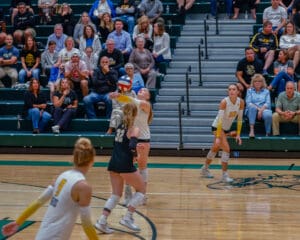 Women's volleyball match, player sets ball while teammates and crowd watch intensely in gymnasium.
