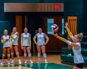 Volleyball player prepares to serve in game, with teammates watching in the background.