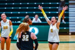 Volleyball team celebrates victory on court, players in white jerseys with numbers 6 and 22, arms raised in excitement.