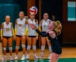 Volleyball player sets ball in action during match, teammates in background wearing uniforms, focused on the game.