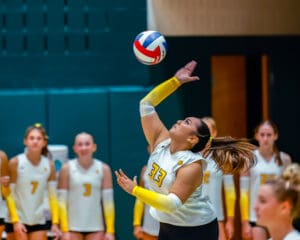 Volleyball player in white jersey serves ball during an indoor match, with teammates watching in the background.