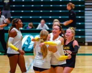 Volleyball team celebrates victory with group hug on indoor court, expressing excitement and camaraderie.
