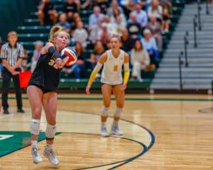 Volleyball player in black uniform performs a pass during a match in a gymnasium with audience in background.