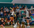 Women's volleyball player spikes ball over net during intense match, with audience watching intently in the background.