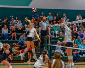 Women's volleyball player spikes ball over net during intense match, with audience watching intently in the background.