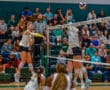 Volleyball player spikes ball over blocker at indoor court, with audience watching in background.