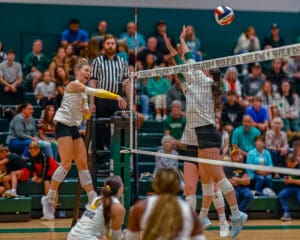Volleyball player spikes ball over blocker at indoor court, with audience watching in background.