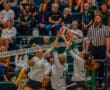 Women's volleyball game, athletes in mid-action at the net with spectators and referee observing.