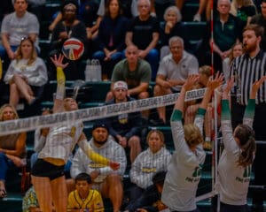 Volleyball game action: player spikes ball over net against two defenders, with audience watching intently in the background.