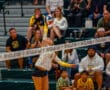 Volleyball player spikes ball over net during an intense match in front of spectators in a gymnasium.