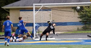 Soccer player scores a goal past the diving goalkeeper during an intense match on a grassy field.