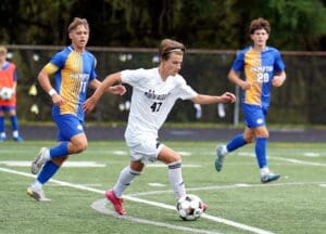Soccer player in white dribbles ball on field, with opposing team players in blue following closely behind.