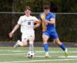 Two soccer players in white and blue jerseys competing for possession on a green field.