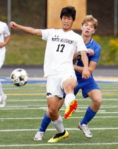 Soccer players in action on the field, one in white jersey kicking the ball, another in blue defending.