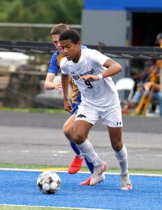 Two soccer players in action on the field, player in white dribbling the ball, opponent close behind.