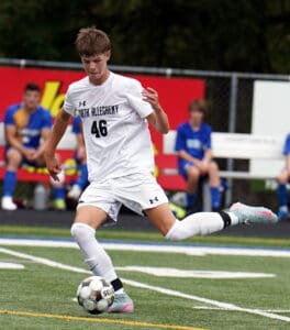 Soccer player in white jersey dribbles ball during a game, with blue-uniformed teammates seated in the background.