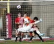 Soccer player in white kicks ball mid-air during match, surrounded by opponents in red jerseys on field.