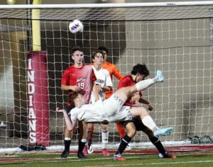 Soccer player in white kicks ball mid-air during match, surrounded by opponents in red jerseys on field.