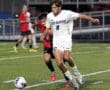 Soccer match action shot: player in white dribbles past opponent in red on a turf field under stadium lights.
