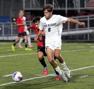Soccer match action shot: player in white dribbles past opponent in red on a turf field under stadium lights.