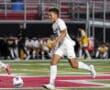 Soccer player dribbles the ball on a field during a match, wearing a white uniform and cleats.