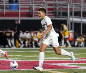 Soccer player dribbles the ball on a field during a match, wearing a white uniform and cleats.