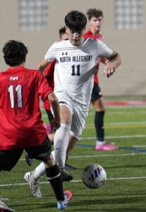 Soccer match action with player in white from North Allegheny dribbling past opponents on field.