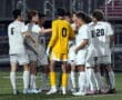 Soccer team huddles on field, players in white and goalie in yellow.