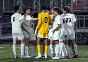 Soccer team huddles on field, players in white and goalie in yellow.