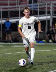 Soccer player from North Allegheny in action on a field at night, wearing white jersey and dribbling a ball.