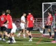 Soccer players mid-game near the goal, team in red defends against team in white during a nighttime match.