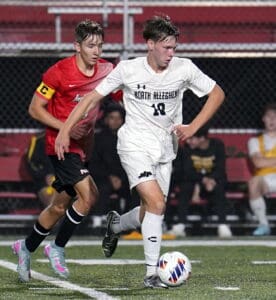 High school soccer players in action, one in white dribbling past an opponent in red on the field.