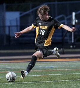 Soccer player in action kicking ball during nighttime match on a field, wearing black and yellow uniform with Tigers logo.