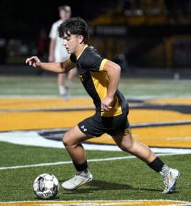 Soccer player in black and yellow uniform dribbling the ball on a field during a night match.