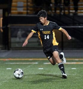 Soccer player in black and yellow uniform, mid-action on the field, kicking the ball under stadium lights.